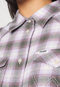Gros plan sur une personne portant une chemise à carreaux violette et verte avec une étiquette logo Wrangler sur la poche poitrine.