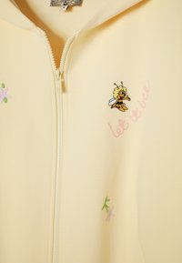 Lysegul lynlåstrøje med lille broderet bi-figur og pink tekst "let it bee" samt små pastelfarvede blomster på stoffet.