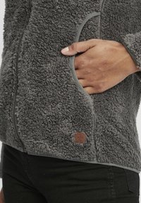 Harmaa fleece-takki, jossa on teksturoitu ja pehmeä pinta. Ominaisuuksiin kuuluu vetoketjukiinnitys, sivutasku ja ruskea logopatchi helman lähellä.