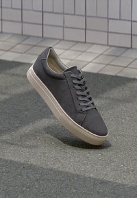 Graue Wildledersneaker mit Gummisohle, flachen Schnürsenkeln, gepolstertem Knöchelbereich und runder zehenpartie, vor einem strukturierten Pflasterhintergrund.