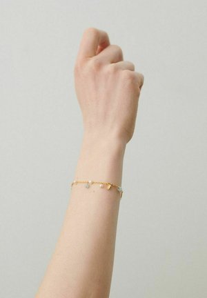 Podlaketnik z nežno zlatim zapestnim trakom z majhnimi obeski in perlicami na enostavni svetli podlagi.