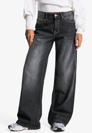 Personne portant un jean noir délavé à jambes larges, un pull côtelé blanc et des chaussures de sport argentées, debout devant un fond blanc.