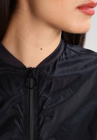 Czarna kurtka bomberka z zamkiem z przodu, ribbowanym kołnierzem i marszczonym materiałem. Posiada mały czarny suwak i gładkie, błyszczące wykończenie.