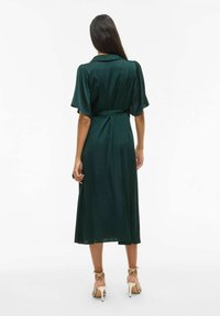 Mørkegrønn sateng midi-kjole med korte puffermærmer, belte i midjen og et flytende skjørt. Sandaler med ankelbånd fullfører looken.