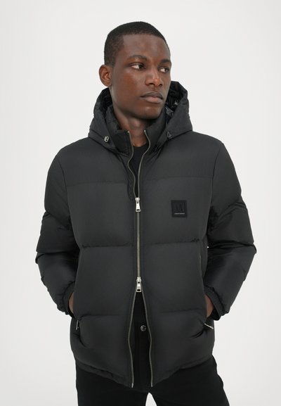 Doudoune noire, avec fermeture éclair à l'avant, capuche, texture matelassée et un petit écusson logo. Coupe slim avec poches latérales.