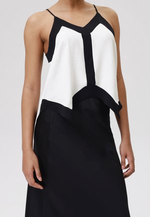 Vrouw draagt een mouwloze top in wit en zwart met dunne bandjes en een zwarte rok tegen een grijze achtergrond.