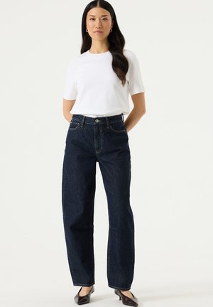 Woman with long dark hair wearing a white short-sleeve shirt, dark blue high-waisted jeans, and dark pointed-toe heels, standing with hands behind back.