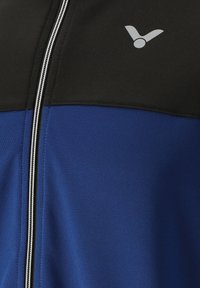 Blå og sort lynlåsjakke lavet af glat stof. Har en kontrasterende øverste sektion og et lille logo på brystet. Sølvlynlås.