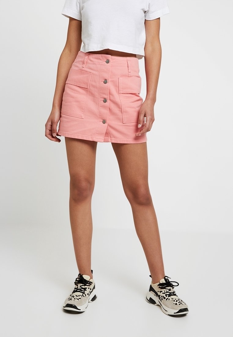 Femme portant une mini-jupe rose à boutons sur le devant avec de grandes poches plaquées, un t-shirt court blanc et des baskets noir et blanc, debout devant un fond blanc.