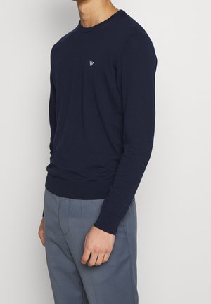 Hombre vistiendo un suéter ajustado de manga larga azul marino con un pequeño logo blanco en el pecho y pantalones grises, posando frente a un fondo sencillo.