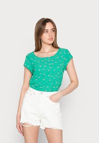 Top verde de mangas curtas com um padrão floral, feito de um tecido leve. Usado com calções de ganga brancos de cintura alta com bordas desfiadas.