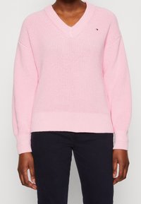 Maglione a V in maglia di un delicato rosa chiaro, con maniche lunghe e polsini a costine. Presenta un piccolo logo del brand sul petto.
