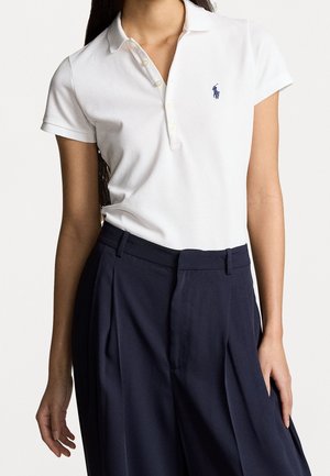 Donna che indossa una polo bianca a maniche corte con un piccolo logo e pantaloni scuri plissettati a vita alta.