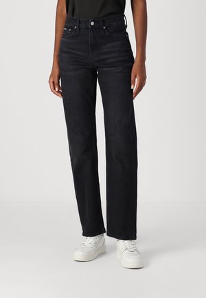 Person iført sorte jeans med højtaljede lige ben kombineret med hvide sneakers og en sort top, stående foran en ensartet baggrund.