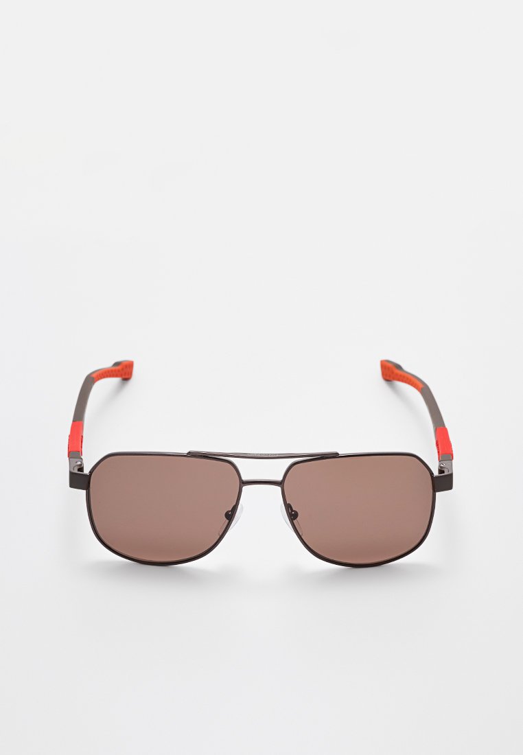 Gafas de sol rectangulares con montura metálica, lentes marrones y patillas negras con empuñaduras de goma rojas sobre un fondo blanco.