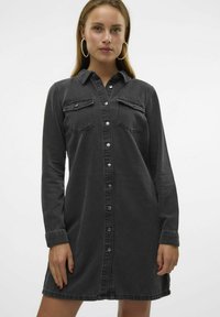 Svart denim skjortekjole med lange ermer, knappestolpe, to brystlommer og buet kant. Har smale mansjetter og lett slitasje.