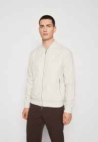 Giacca bomber beige chiaro in un morbido tessuto testurizzato con zip frontale, polsini e vita a coste, indossata sopra pantaloni scuri.