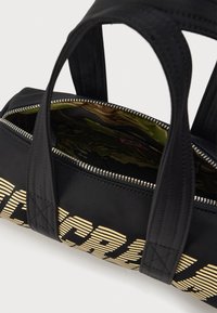 Μαύρη τσάντα με εμφανές κίτρινο ριγέ λογότυπο. Διαθέτει διπλές λαβές, κλείσιμο με φερμουάρ και εσωτερική επένδυση με πράσινο σχέδιο.