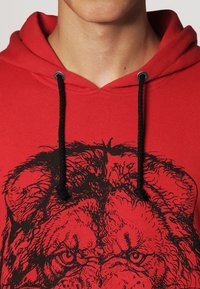 Roter Kapuzenpullover mit schwarzem Löwengrafikdruck auf der Brust. Verfügt über Kordelzüge, Metallösen und einen weichen, strukturierten Stoff.