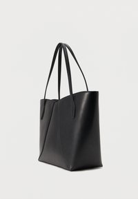 Sac cabas en cuir noir avec deux longues bandoulières et un panneau latéral légèrement incliné sur un fond clair uni.