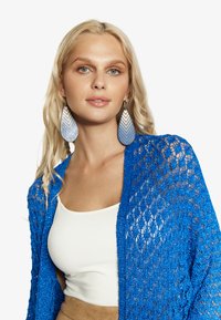 Elektriski zila mežģīņu kardigāns virs baltā topa; modele valkā lielas zilas gradienta auskarus ar ševronu rakstu, redzama maiga tekstūra.