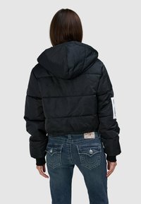 Czarna, krótka kurtka puchowa z kapturem, posiadająca pikowany wzór i elastyczne mankiety, zestawiona z ciemnoniebieskimi dżinsami z ozdobnymi tylkimi kieszeniami.