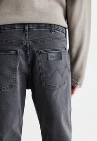 Pilkos džinsai su Wrangler logotipo pleistru dešinėje nugarinėje kišenėje, pasižymintys kontrastuojančia siūle ir laisvu siluetu.