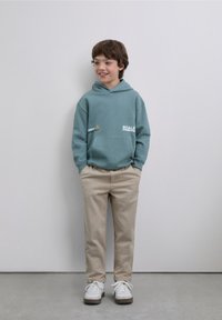 Turkooise hoodie met een kangoeroezak en tekstgrafiek, gecombineerd met beige broek en witte sneakers, tegen een effen achtergrond.