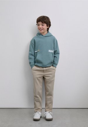 Sudadera con capucha color verde azulado con bolsillo tipo canguro y gráfico de texto, combinada con pantalones beige y zapatillas blancas, sobre un fondo liso.