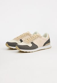 Zapatillas deportivas en color beige y gris con un upper texturizado, una media suela blanca y cordones de color claro. Presentan un logo en el lateral.