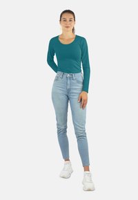 Türkiser Langarmoberteil mit Rundhalsausschnitt, kombiniert mit hochgeschnittenen hellblauen Jeans. Das Model trägt weiße Sneaker und einen gelben Ohrring.
