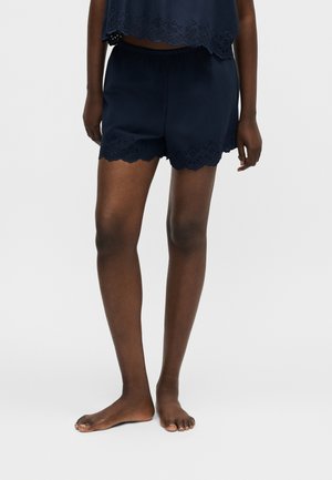 Személy sötétkék hullámos szélű rövidnadrágot és hozzáillő felsőt visel, mezítláb áll egy egyszínű világos háttéren.