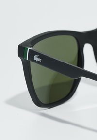 Fekete napszemüveg zöld árnyalatú lencsékkel. A kereten zöld és fehér csík található, valamint egy ezüst krokodil logó a szárán.