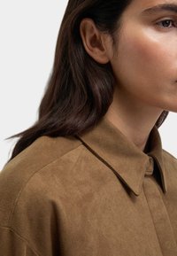 Chemise en daim marron avec un col, présentant une texture douce et une fermeture à boutons. Gros plan sur le torse supérieur et le profil latéral.