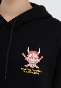 Haut à capuche noir avec un graphique rouge et blanc représentant un visage de démon avec des cornes, des épées croisées en dessous, et le texte "FACES DES SANS VISAGE" en jaune.