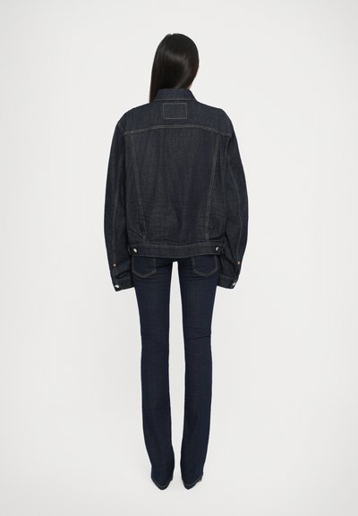 Veste en denim foncé à la coupe décontractée, avec des coutures visibles et des accents de boutons. Associée à un jean foncé évasé, vue de dos.