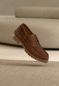 Καφέ δερμάτινο παπούτσι τύπου boat με κορδόνια και ανθεκτική σόλα, εμφανιζόμενο στον αέρα πάνω από μπεζ χαλί και φόντο με καναπέ.