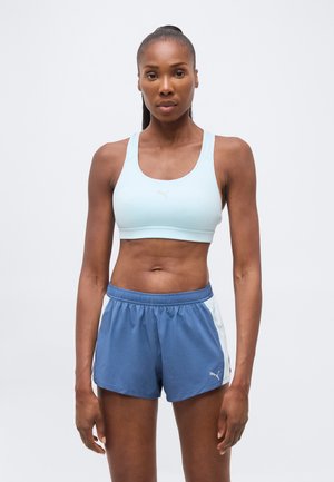 Femeie cu piele închisă la culoare și păr prins la spate, purtând un sutien sport albastru deschis și pantaloni scurți sport Puma albaștri, stând pe un fundal simplu.