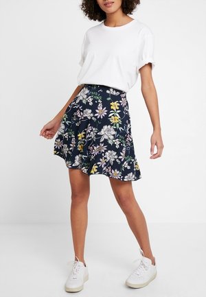 Femme portant un t-shirt blanc, une jupe fleurie bleu marine et des baskets blanches, debout devant un fond blanc uni.