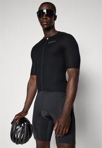Svart sykkeltrøye med korte ermer og frontglidelås, kombinert med svarte polstrede shorts. Modellen holder en svart sykkelhjelm.