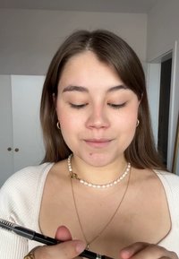 Giovane che tiene una matita per sopracciglia nera con un pennello a spirale. Indossa un maglione bianco e una collana di perle con un gancio d'oro.