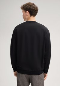 Felpa nera con scollatura a girocollo e polsini a coste. Vestibilità ampia, texture liscia, design semplice, indossata con pantaloni grigi.