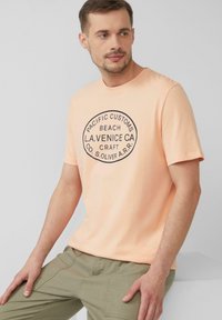 Perzikkleurig t-shirt met korte mouwen, voorzien van een cirkelvormige zwarte grafiek met tekst. Gemaakt van zacht materiaal, met een ronde hals en een relaxte pasvorm.