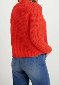 Pomarańczowy sweter dzianinowy z teksturowaną, prążkowaną powierzchnią. Posiada luźny krój i okrągły dekolt, w połączeniu z niebieskimi dżinsami.