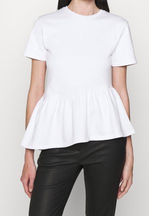 Femme portant un top péplum blanc à manches courtes associé à un pantalon noir ajusté, présenté du cou jusqu’aux hanches sur un fond uni.