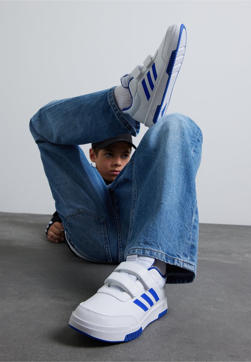 Baskets blanches avec des accents bleus, dotées de deux bandes Velcro, d'une semelle texturée et d'un design sportif, associées à un pantalon en denim bleu.