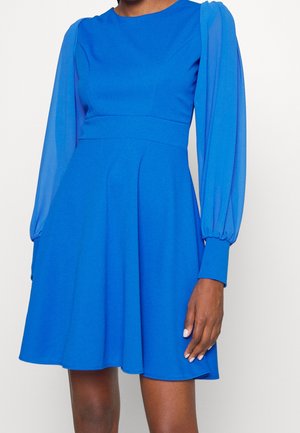 Woman wearing a bright blue knee-length dress with long sheer puff sleeves and fitted waist.