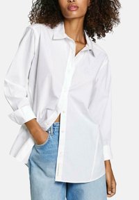 Camicia bianca con bottoni e maniche lunghe, caratterizzata da un colletto a punta, taglio dritto e leggera texture. Abbinata a jeans in denim azzurro chiaro.