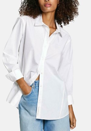 Camicia bianca con bottoni e maniche lunghe, caratterizzata da un colletto a punta, taglio dritto e leggera texture. Abbinata a jeans in denim azzurro chiaro.
