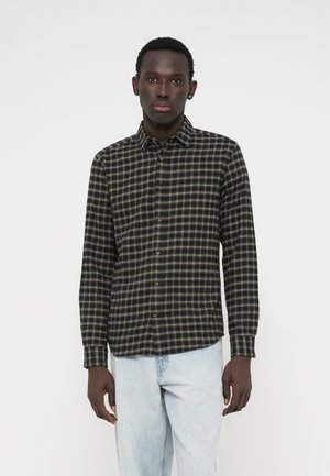 Giovane uomo con pelle scura che indossa una camicia a maniche lunghe a scacchi nera e beige e jeans azzurri chiari, in piedi di fronte a uno sfondo bianco semplice.